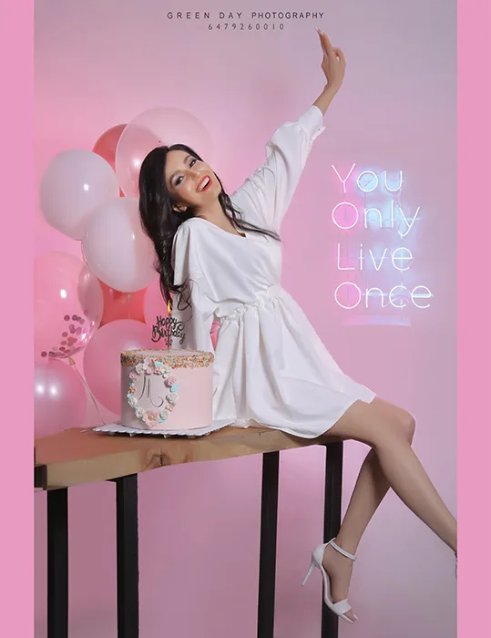 Woman in a white outfit seated beside a birthday cake with pink balloons and one arm raised