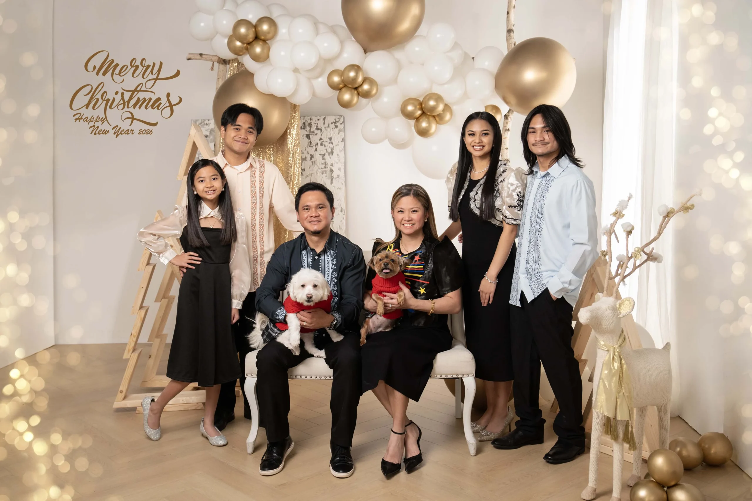Family posing with two dogs in red sweaters against a cream and gold Christmas background