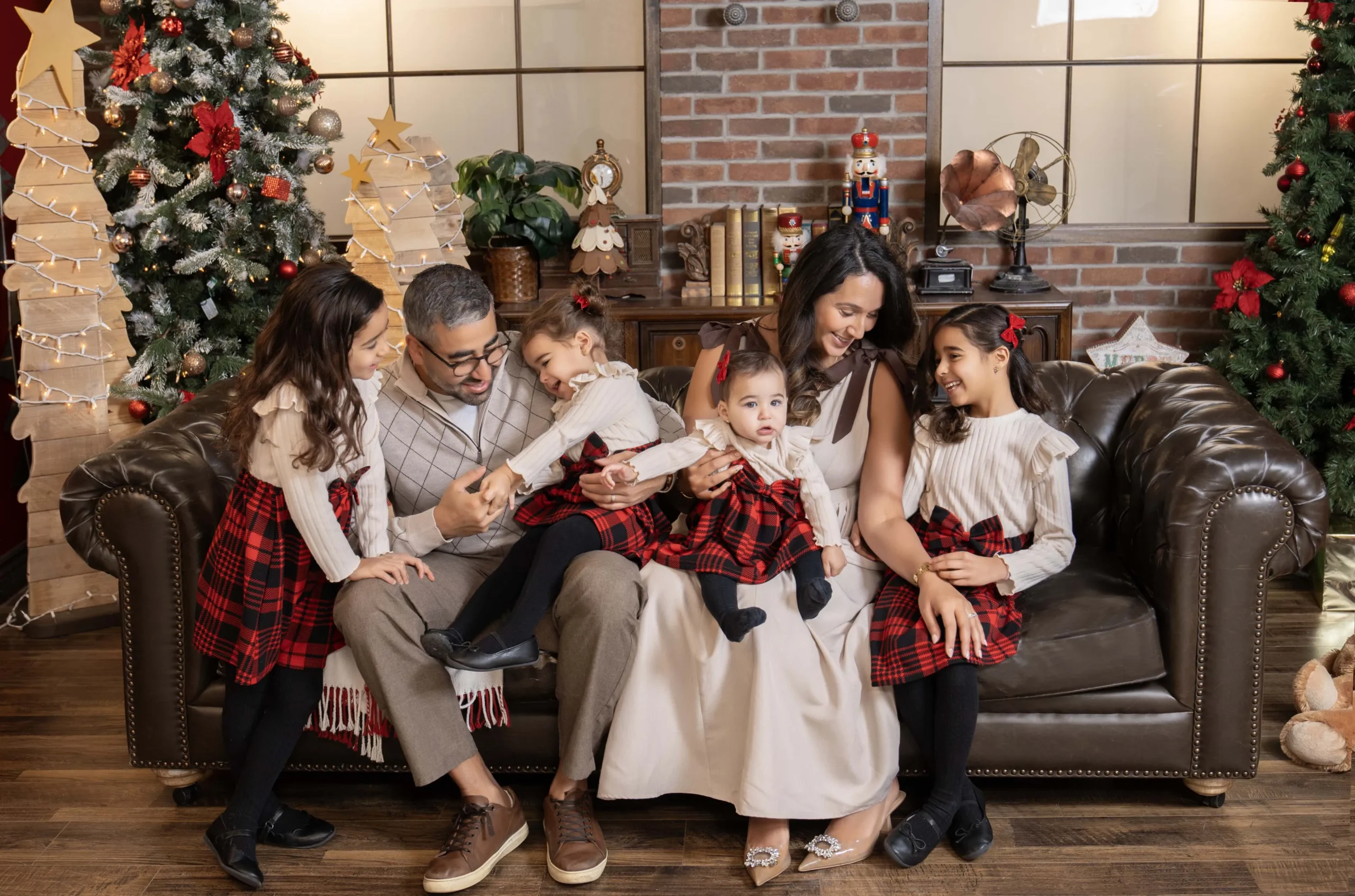 Parents sharing a playful moment with four children on a brown sofa surrounded by Christmas trees