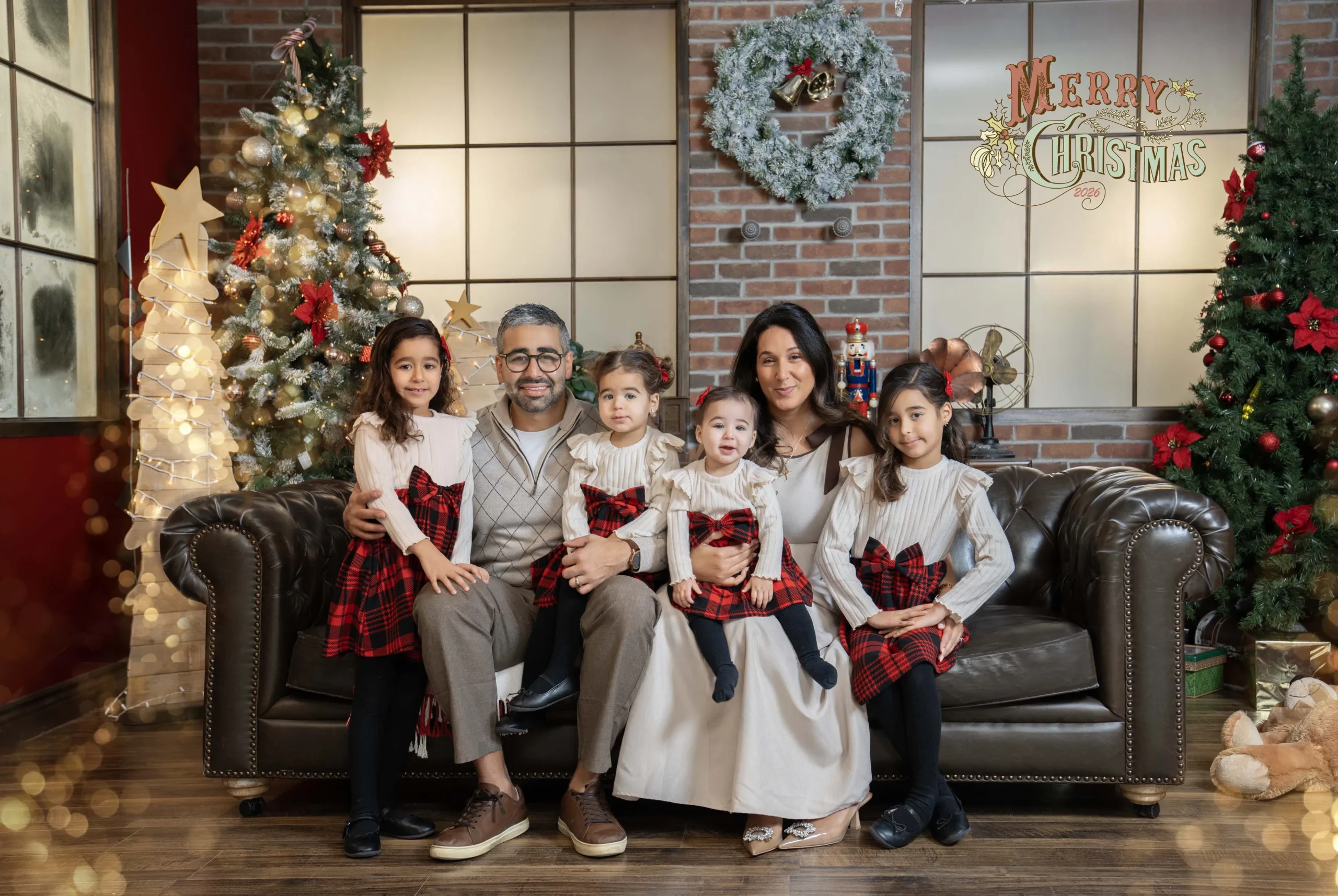 Parents and four children in cream and red plaid outfits posing on a brown sofa beside decorated Christmas trees