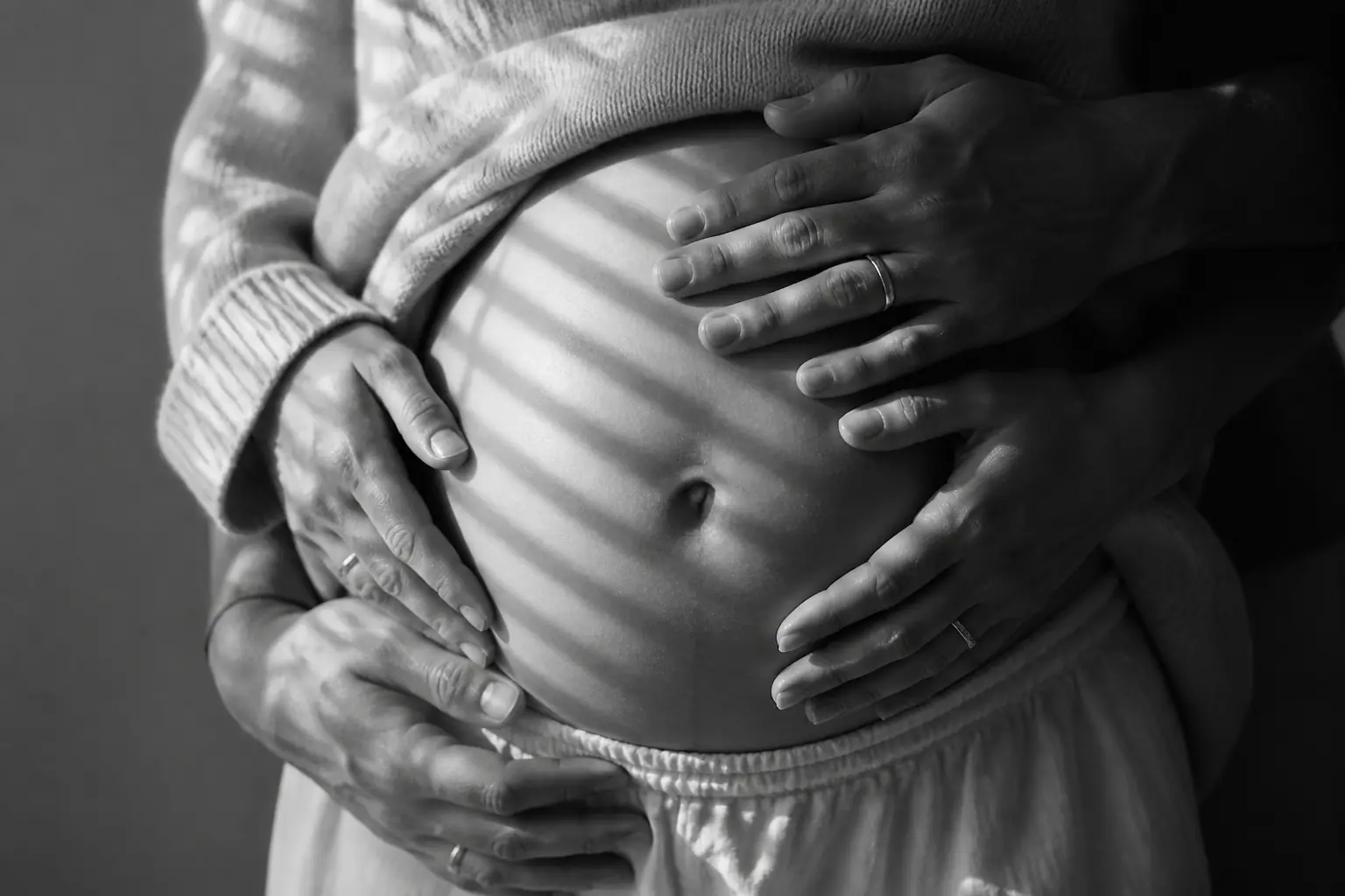 Black-and-white close-up of an expecting couple's hands around a pregnant belly