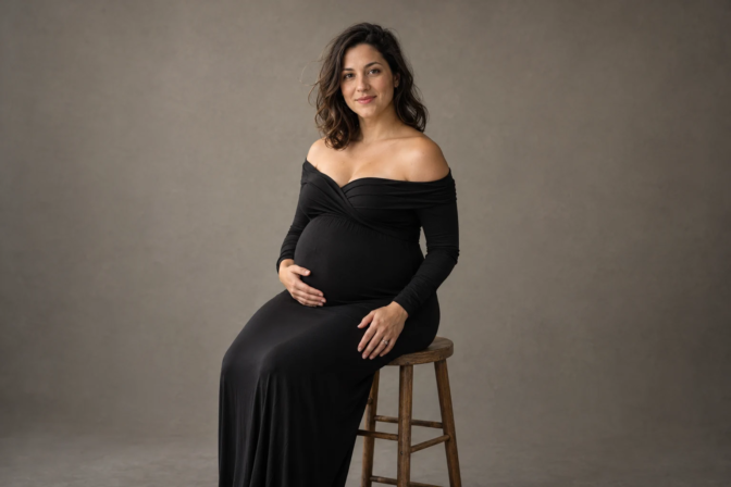 Pregnant woman in a black off-shoulder dress seated on a wooden stool