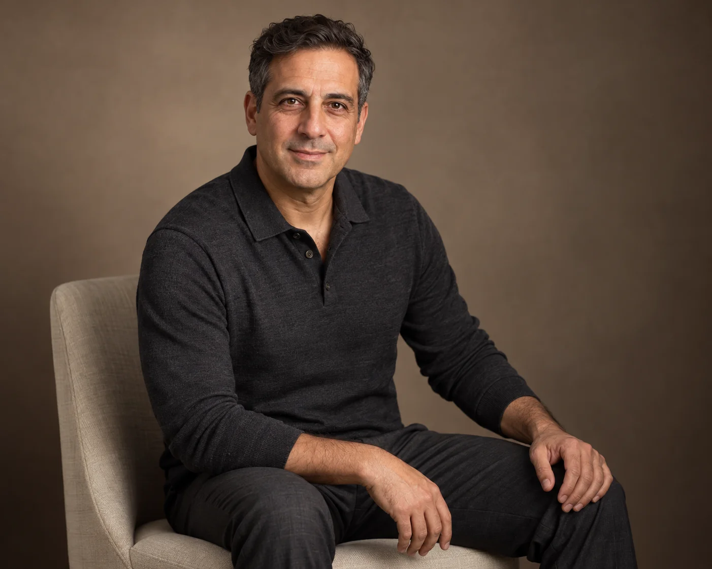 Illustrated relaxed business portrait of a seated man in a charcoal polo against a warm neutral studio background
